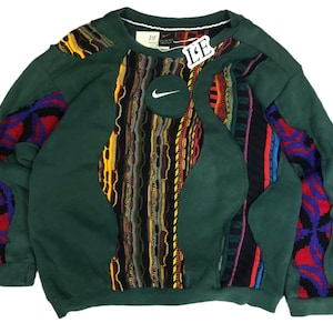 May include: A green sweatshirt with a unique patchwork design. The center features a textured pattern with yellow, red, and black threads. The Nike logo is on a circular patch. Sleeves have colorful geometric patterns.