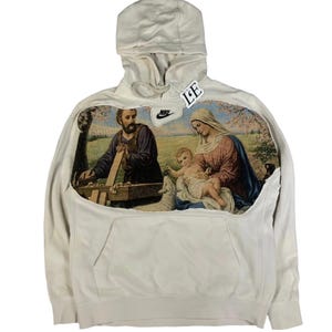 Op de afbeelding: Crèmekleurige hoodie met een religieus tapijtdesign. Het tapijt toont een bijbelse scène met figuren in gewaden. Een Nike-logopatch is zichtbaar. De hoodie heeft een capuchon en een voorzak.