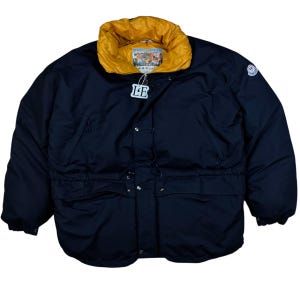 Puede incluir: Una chaqueta acolchada azul marino con cuello amarillo brillante. La chaqueta tiene cintura con cordón, cierres a presión y un pequeño parche con el logotipo en la manga. Es visible una pequeña etiqueta con las letras "LE".