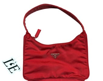 May include: A red Prada handbag with a curved shape and a single handle. The bag features a zipper closure and a small silver Prada logo. A white tag with the letters "L" and "E" is attached to the bag.