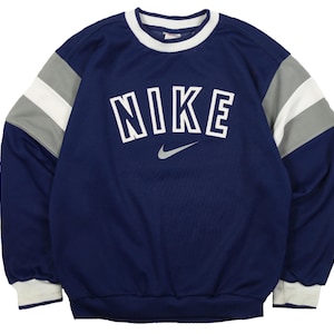Op de afbeelding: Marineblauwe Nike-trui met witte, grijze en marineblauwe accenten. Het woord "NIKE" is in witte blokletters op de borst gedrukt, met daaronder een grijs Nike swoosh-logo. De mouwen hebben grijze en witte strepen.