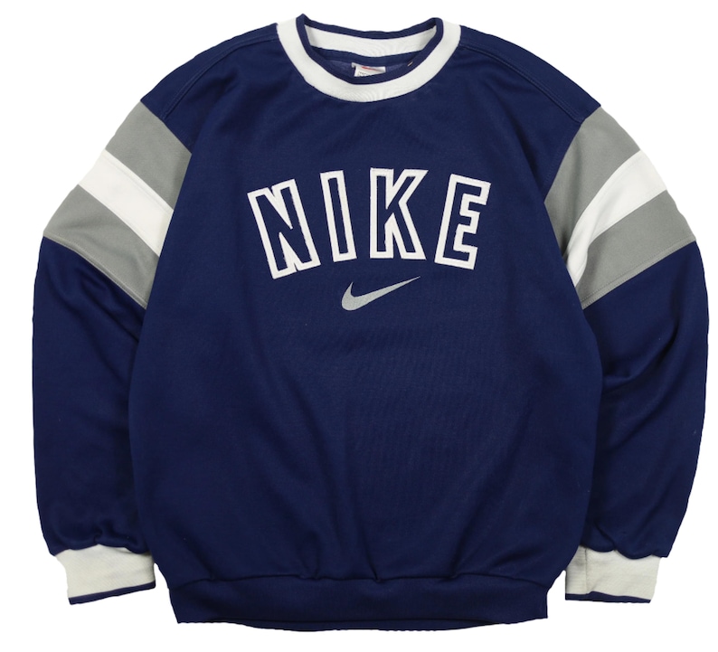 Op de afbeelding: Marineblauwe Nike-trui met witte, grijze en marineblauwe accenten. Het woord "NIKE" is in witte blokletters op de borst gedrukt, met daaronder een grijs Nike swoosh-logo. De mouwen hebben grijze en witte strepen.