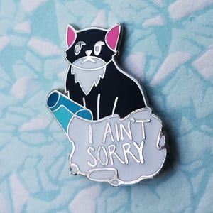 May include: Black cat enamel pin with pink ears and a blue and white striped object. The pin says "I ain't sorry".