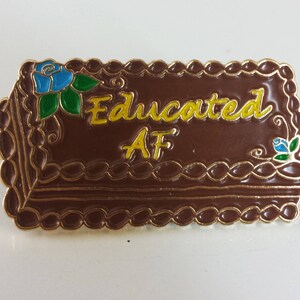 May include: A brown and gold enamel pin shaped like a cake with the words "Educated AF" in yellow lettering and blue and green floral accents.