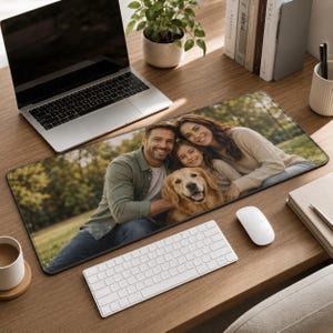 May include: A rectangular desk mat featuring a printed photograph of a family consisting of a man, a woman, and a young child posing outdoors with a golden retriever dog. The mat is positioned on a wooden desk surface beneath a laptop and keyboard.