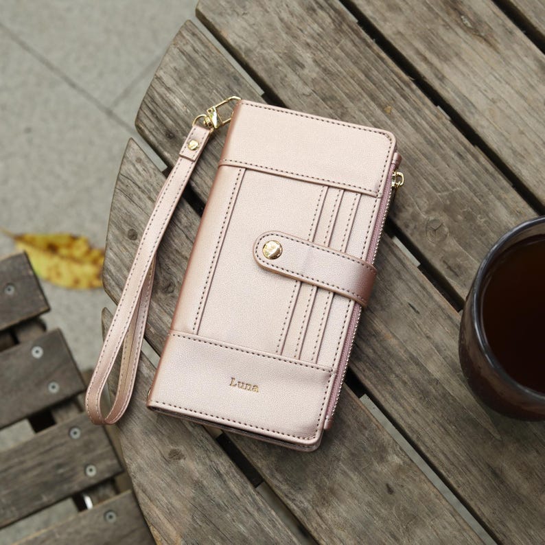 May include: A rose gold wallet with a wrist strap, resting on a wooden surface. The wallet features a snap closure, multiple card slots, and the name "Luna" embossed on the front. The strap and zipper pull are gold-toned.