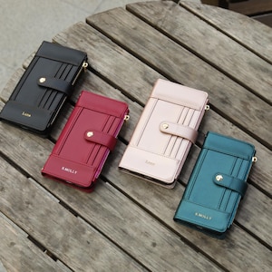 May include: Four leather wallets in black, burgundy, blush pink, and teal, arranged on a wooden surface. Each wallet has a snap closure, card slots, and a zipper. The black and pink wallets have the word "Lune" printed on them, while the burgundy and teal wallets have "S.MOLLY".