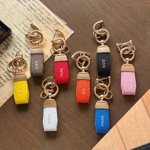 Pode incluir: Uma coleção de chaveiros de couro em várias cores, incluindo amarelo, vermelho, laranja, preto, rosa, branco, azul e castanho. Cada chaveiro apresenta um anel e fecho de metal dourado, com texto em relevo na etiqueta de couro. O texto inclui nomes e iniciais.
