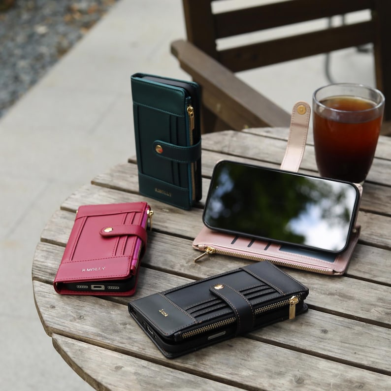 May include: A collection of leather phone cases in various colors, including black, burgundy, and green, with gold zippers and clasps. The cases are arranged on a wooden table, showcasing their design and functionality. A smartphone rests on a pink case.