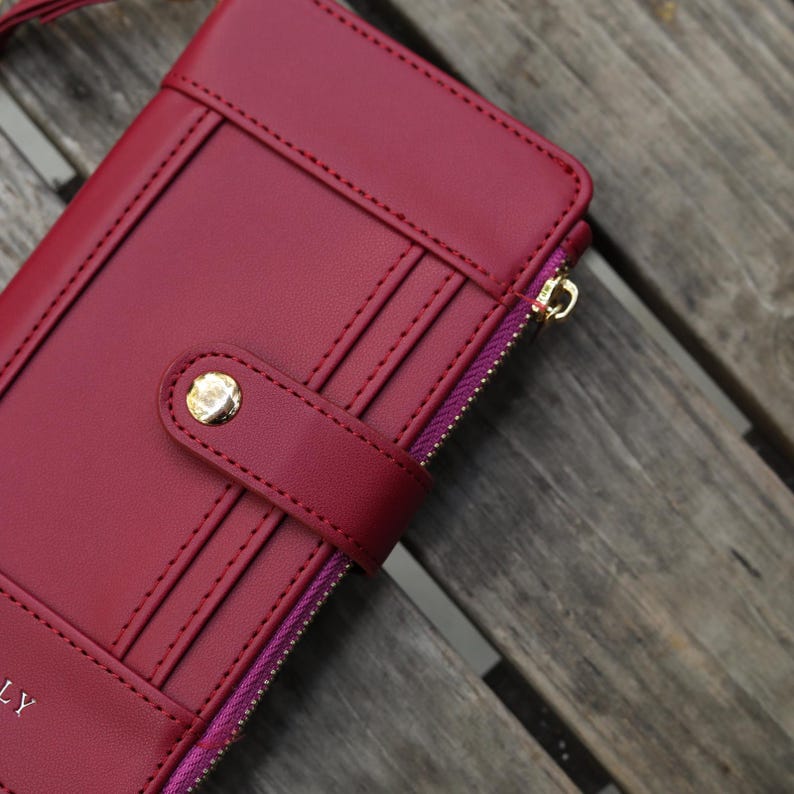 May include: A burgundy leather wallet with a gold-colored zipper pull and snap closure. The wallet has multiple card slots and a stitched design. The zipper is purple. The wallet is on a wooden surface.