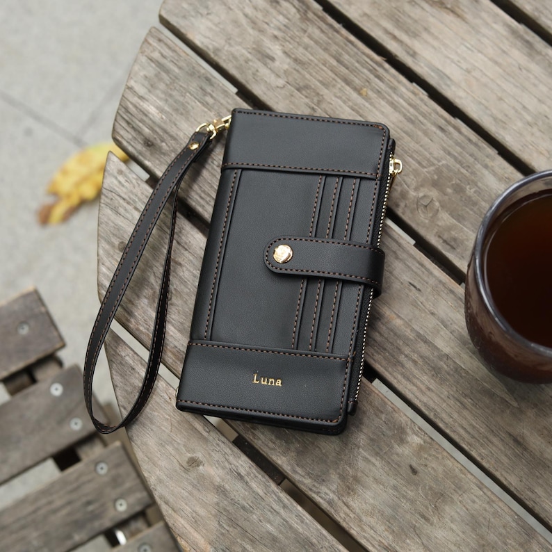May include: Black leather wallet with a wrist strap, featuring a snap closure and multiple card slots. The wallet has a gold zipper and the name "Luna" is printed on the front. The wallet is on a wooden table.