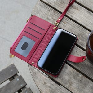 May include: Open burgundy leather wallet-style phone case with a wrist strap. The wallet has a zipper pocket, card slots, and a clear ID window. A black smartphone is secured inside the case, with a burgundy strap closure.