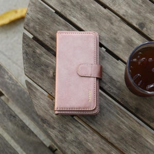 May include: A rose gold phone case with a magnetic closure and the name "SARAH" in gold lettering. The case is made of a smooth, leather-like material. A glass of iced tea sits nearby on a wooden table.