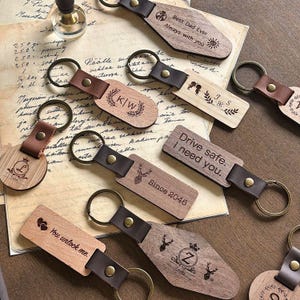 Puede incluir: Llaveros de madera con diseños grabados y texto. Los llaveros tienen correas de cuero y anillos de metal. Algunos llaveros tienen iniciales, ciervos o el texto "Best Dad Ever" y "Drive safe. I need you."