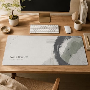 Puede incluir: Una alfombrilla de escritorio rectangular con fondo gris claro, una gran pincelada circular gris oscuro, acentos de acuarela verde salvia y líneas horizontales finas. El texto Noah Bennett y Create with Calm está impreso en negro.