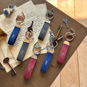 Peut inclure: Plusieurs porte-clés personnalisés de différentes couleurs, dont le bleu, le gris et le bordeaux. Chaque porte-clés est doté d'une lanière en cuir avec des initiales ou un nom gravé. Les porte-clés sont fixés à des anneaux métalliques.