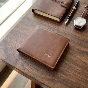 Puede incluir: Una cartera de cuero marrón con el texto en relieve "L. Kibler" se muestra sobre una superficie de madera. También se ven un cuaderno de cuero a juego, un bolígrafo y un reloj de pulsera con correa de cuero marrón.