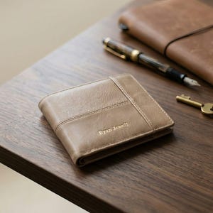 Puede incluir: Una cartera de cuero marrón claro con el nombre "Ryan Jewell" grabado en la parte delantera. La cartera es rectangular y tiene un diseño cosido. Un bolígrafo, una llave y un cuaderno de cuero también están sobre la superficie de madera.