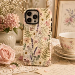 May include: A smartphone case featuring a cream-colored background with a detailed botanical print of pressed wildflowers, including purple lavender sprigs, green fern leaves, white daisies, and small pink blossoms, with a black camera cutout.