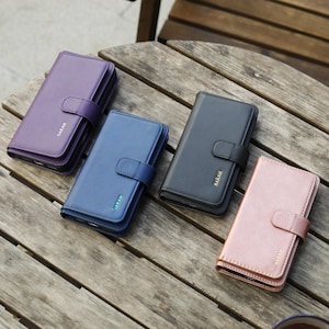 May include: Four leather phone cases in purple, blue, black, and rose gold, each with a matching clasp and the name "SARAH" embossed on the front. The cases are arranged on a wooden table.