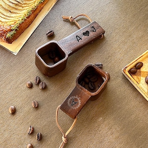 May include: Two wooden coffee scoops with leather handles, filled with coffee beans. One scoop has the initials "A" and "J" with a heart symbol. The other scoop has the initial "A" with a decorative design. Coffee beans are scattered around.