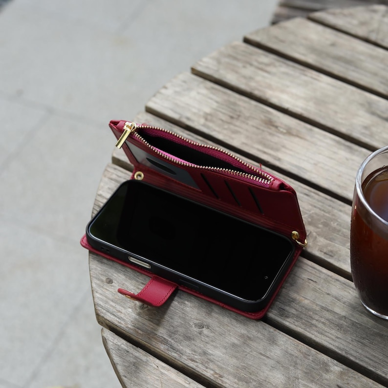 May include: A red leather phone case with a gold zipper and multiple card slots. The case is open, revealing a black smartphone. The case is on a wooden table next to a glass of iced tea.