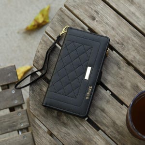 Könnte beinhalten: Schwarze Geldbörse mit einem gesteppten Rautenmuster und einem goldfarbenen Metallakzent. Die Geldbörse hat eine Handschlaufe und den Text "S.GRACE" in Gold. Die Geldbörse liegt auf einem Holztisch.