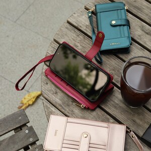 May include: A collection of leather phone cases and wallets in various colors, including teal, burgundy, and rose gold, arranged on a wooden surface. One case holds a smartphone. A glass of dark liquid sits nearby.