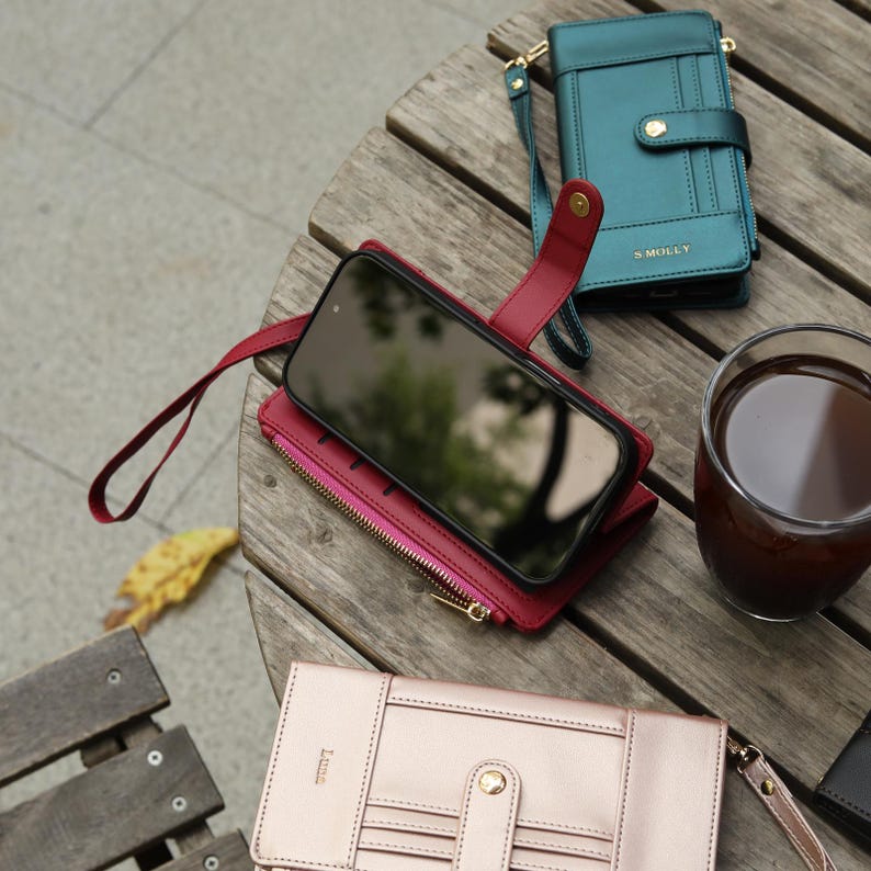 May include: A collection of leather phone cases and wallets in various colors, including teal, burgundy, and rose gold, arranged on a wooden surface. One case holds a smartphone. A glass of dark liquid sits nearby.