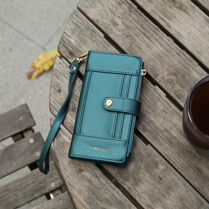 May include: Teal wallet with a wrist strap, resting on a wooden surface. The wallet has a snap closure and a zipper. The text "S Molly" is visible on the front. A cup of tea is in the background.