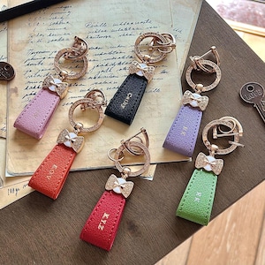 Pode incluir: Uma coleção de chaveiros com etiquetas de couro em várias cores: rosa, preto, roxo, vermelho e verde. Cada chaveiro apresenta um anel dourado adornado com strass e um pingente em forma de laço. As etiquetas têm iniciais.