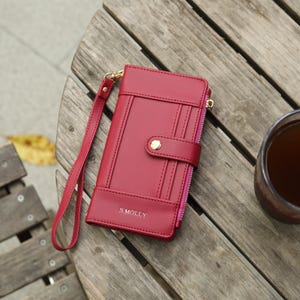 May include: A red wallet with a wrist strap, resting on a wooden surface. The wallet has a snap closure and a zipper. The text "S. MOLLY" is visible on the front. A cup of coffee sits nearby.
