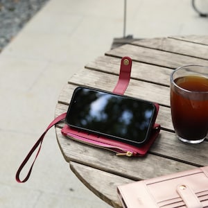 May include: A red leather phone case with a zipper pocket and wrist strap holds a black smartphone. A glass of dark liquid sits next to the case on a wooden table. A second pink wallet is also on the table.