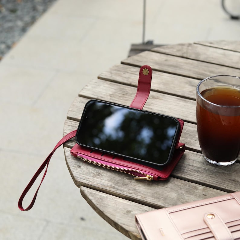 May include: A red leather phone case with a zipper pocket and wrist strap holds a black smartphone. A glass of dark liquid sits next to the case on a wooden table. A second pink wallet is also on the table.