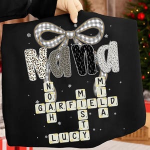 May include: Black tote bag with the word "NANA" in a mix of patterns and a bow at the top. Below, a Scrabble-style design spells out "GARFIELD", "LUCY", and "MIA". The bag is held up by a person.