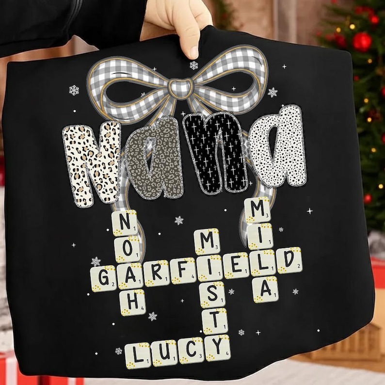 May include: Black tote bag with the word "NANA" in a mix of patterns and a bow at the top. Below, a Scrabble-style design spells out "GARFIELD", "LUCY", and "MIA". The bag is held up by a person.
