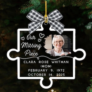 May include: A clear acrylic puzzle piece ornament with a photo of a woman, the text "Our Missing Piece," and the name Clara Rose Whitman, with birth and death dates. It is topped with a black and white checkered bow.