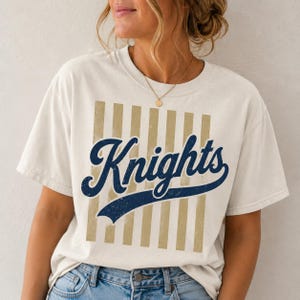 May include: A cream-colored short-sleeve t-shirt featuring a graphic design with vertical tan stripes and the word Knights written in a dark blue cursive script font across the center. The shirt has a crew neckline and a relaxed fit.
