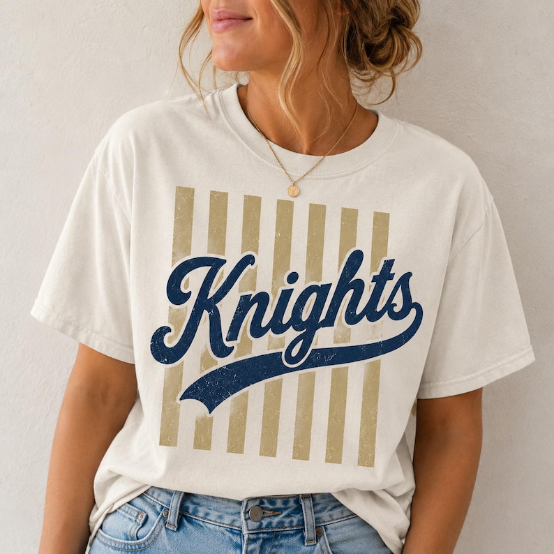 May include: A cream-colored short-sleeve t-shirt featuring a graphic design with vertical tan stripes and the word Knights written in a dark blue cursive script font across the center. The shirt has a crew neckline and a relaxed fit.
