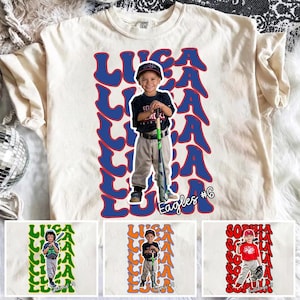 Puede incluir: Camiseta color crema con una foto de un niño con uniforme de béisbol, con el nombre "LUCA" en letras azules de burbujas. También es visible el texto "Eagles #6". La camiseta es una prenda informal.