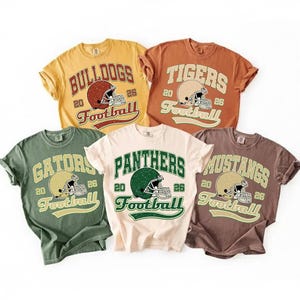 Puede incluir: Cinco camisetas de estilo vintage con diseños de fútbol americano. Cada camiseta presenta el nombre de un equipo, un gráfico de casco de fútbol y la palabra "Football". Las camisetas son de varios colores: amarillo, naranja, verde, crema y marrón.