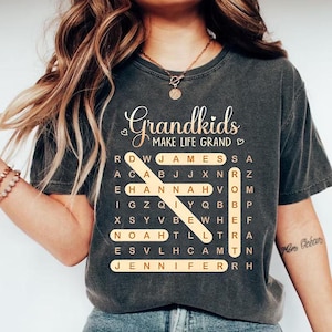 May include: Dark gray t-shirt with the words "Grandkids make life grand" in a cursive font. A word search grid includes names like James, Hannah, and Jennifer. The shirt is a comfortable, casual style.