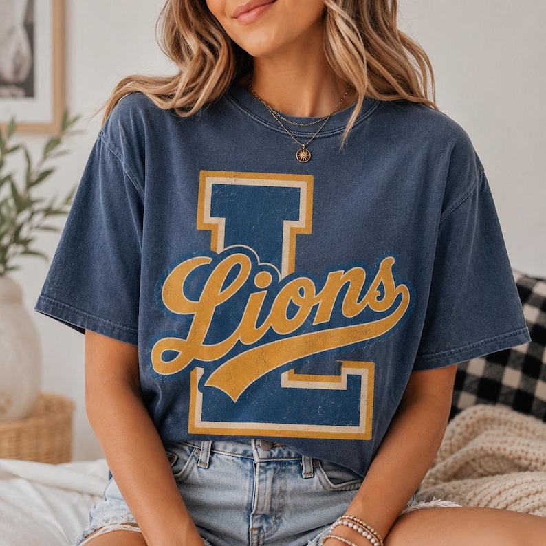 May include: A faded navy blue short-sleeve t-shirt featuring a large block letter L in blue with a gold outline, overlaid with the word Lions in a gold cursive script font. The shirt has a crew neckline and a relaxed, vintage-style fit.