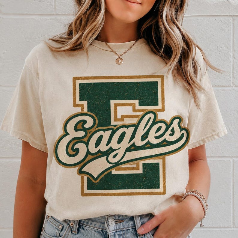 May include: A cream-colored short-sleeve t-shirt featuring a large, distressed dark green block letter E with a gold outline. Overlaid on the letter is the word Eagles in a cursive, dark green font with a gold outline, creating a vintage look.
