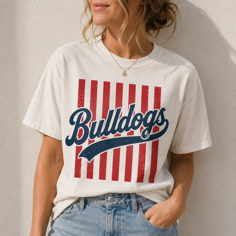 May include: A cream-colored short-sleeve t-shirt featuring a graphic design with vertical red stripes and the word Bulldogs written in a dark blue cursive script font across the center. The shirt has a crew neckline and a relaxed fit.