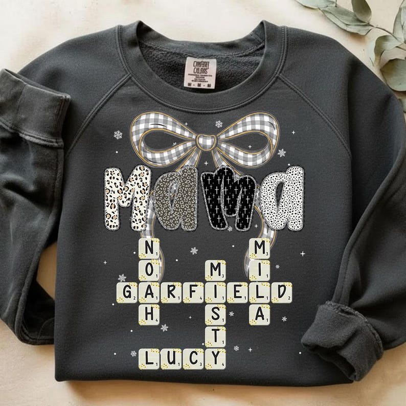 May include: Dark gray sweatshirt with the word "Mama" in a decorative font, with a plaid bow and snowflake accents. Below, names are spelled out in a crossword puzzle style. The sweatshirt is a Comfort Colors brand.