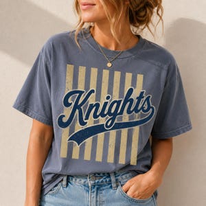 May include: A slate blue short-sleeve t-shirt featuring a distressed graphic print. The design includes vertical beige stripes behind the dark blue script text that reads Knights, with a curved underline extending from the final letter of the word.