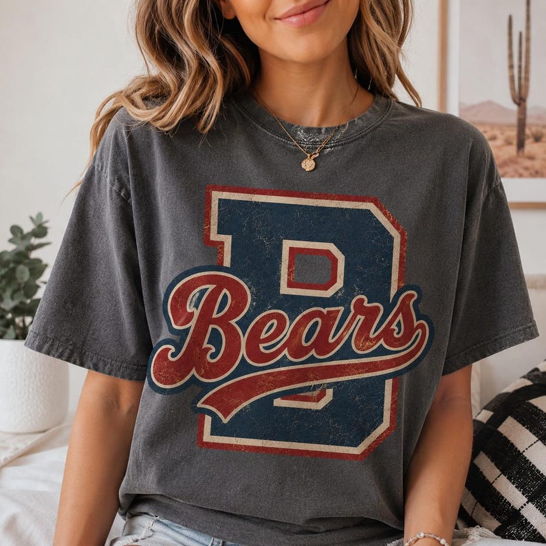 May include: A charcoal gray short-sleeve t-shirt featuring a large, distressed graphic on the front. The design includes a bold, navy blue block letter B layered with the word Bears in a red, cursive script with a white outline.