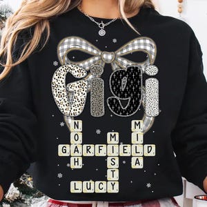 May include: Black sweatshirt featuring the name "Gigi" in a mix of patterns, including leopard print and crosses, above a crossword-style arrangement of names. A large plaid bow is at the top. The names include Garfield, Lucy, and others.