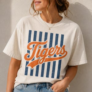 May include: A cream-colored short-sleeve t-shirt featuring a graphic design with vertical navy blue stripes and the word Tigers written in a large, distressed orange script font across the center. The shirt has a crew neckline and a relaxed fit.